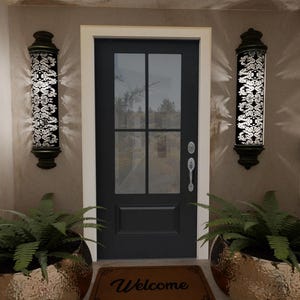May include: A black front door with glass panels is flanked by two decorative wall sconces with intricate floral patterns. Below, two large gold planters hold green ferns, and a welcome mat rests on the floor.