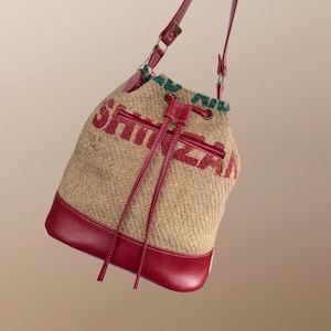 May include: A tan burlap bucket bag with a red leather base and drawstring closure. The bag has a red leather shoulder strap and the word "SHINZA" printed in red across the front. The top edge has green accents.
