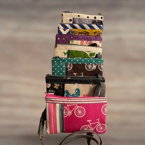 May include: A stack of nine colorful fabric purses with different patterns, including bicycles, birds, and geometric designs. The purses are all small and have a strap.