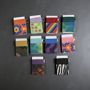 May include: A collection of ten small, rectangular wallets with various colorful patterns. Each wallet has a white top, a patterned front, and a black or colored elastic band. Some designs include rainbows, polka dots, and geometric shapes. Each wallet has a small metal ring.