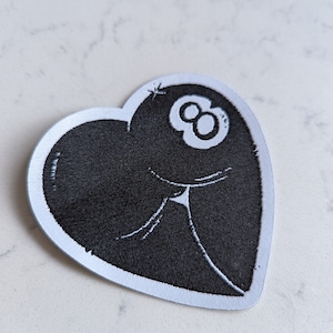 May include: A black and white embroidered patch in the shape of a heart with a white 8-ball inside.