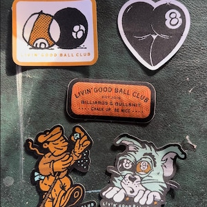 May include: A collection of stickers with a billiards theme. Designs include a black eight ball, a heart with an eight ball, and cartoon characters. The stickers feature the text "LIVIN' GOOD BALL CLUB" and "BILLIARDS & BULLSHIT".