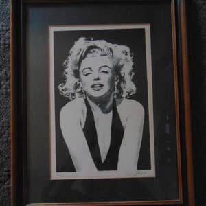 May include: A black and white framed print of a woman with blonde hair. The woman is wearing a black dress and has a serious expression on her face. The print is signed in the bottom right corner.