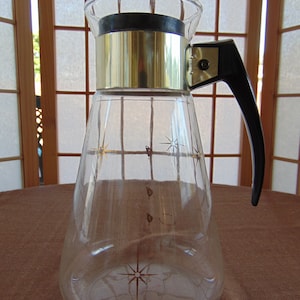 May include: A clear glass pitcher with a silver band around the top and a black handle. The pitcher has a starburst design on the bottom and a few stars on the sides.