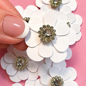 May include: A collection of layered white paper flowers, each featuring a central circular embellishment made of a large clear rhinestone surrounded by a ring of smaller clear crystals set in a gold-toned metal frame, arranged in a cluster.