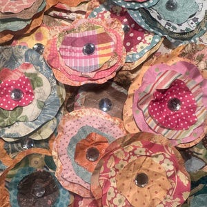 May include: A collection of handmade paper flowers in various colors and patterns. Each flower features multiple layers of petals and a sparkling gem center. The flowers are made from patterned paper in shades of pink, orange, blue, and brown.