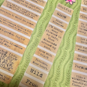 Puede incluir: Primer plano de varias tiras de papel con citas y frases inspiradoras en fuente de m&aacute;quina de escribir. Las tiras son de color beige con rayas horizontales blancas. Una peque&ntilde;a flor de papel rosa y morado est&aacute; en el fondo. El tema es el crecimiento y la naturaleza.