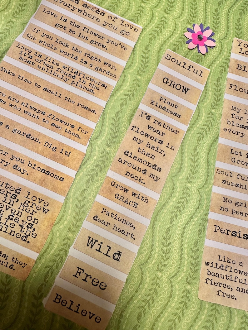 Puede incluir: Primer plano de varias tiras de papel con citas y frases inspiradoras en fuente de m&aacute;quina de escribir. Las tiras son de color beige con rayas horizontales blancas. Una peque&ntilde;a flor de papel rosa y morado est&aacute; en el fondo. El tema es el crecimiento y la naturaleza.