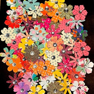 May include: A collection of colorful paper flowers with glitter and rhinestones. The flowers are arranged in a cluster and have a variety of patterns and designs.