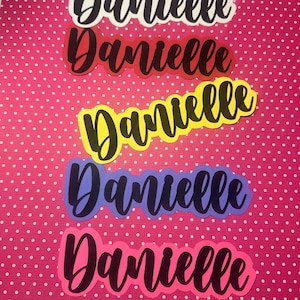 May include: Five colorful cut-out name tags spelling "Danielle" in a cursive font. The tags are white, red, yellow, purple, and pink, each with a black outline. The background is a pink polka dot pattern.