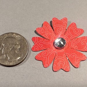 May include: A red paper flower with a white swirl pattern and a silver rhinestone in the center. The flower is about the size of a quarter.