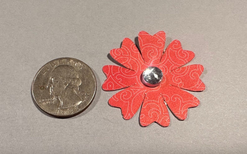 May include: A red paper flower with a white swirl pattern and a silver rhinestone in the center. The flower is about the size of a quarter.