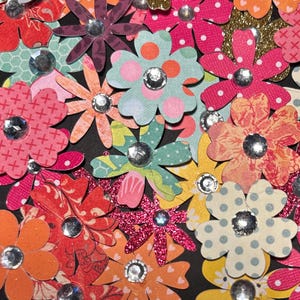 May include: A collection of colorful paper flowers with glitter and rhinestones. The flowers are arranged in a random pattern on a black background.