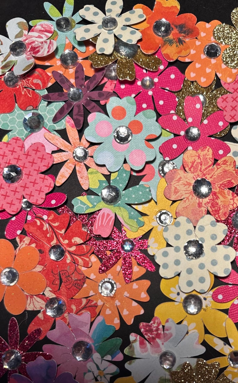 May include: A collection of colorful paper flowers with glitter and rhinestones. The flowers are arranged in a random pattern on a black background.