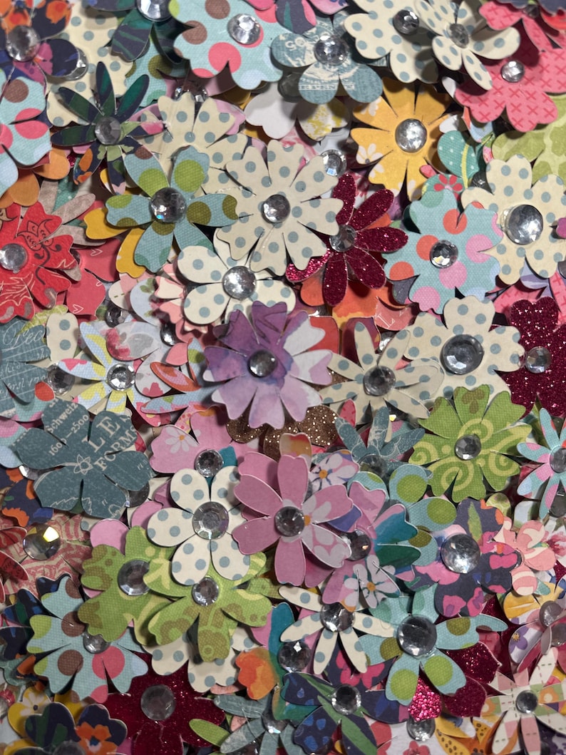 May include: A collection of colorful paper flowers with glitter and rhinestones. The flowers are in various shades of pink, blue, green, and purple.