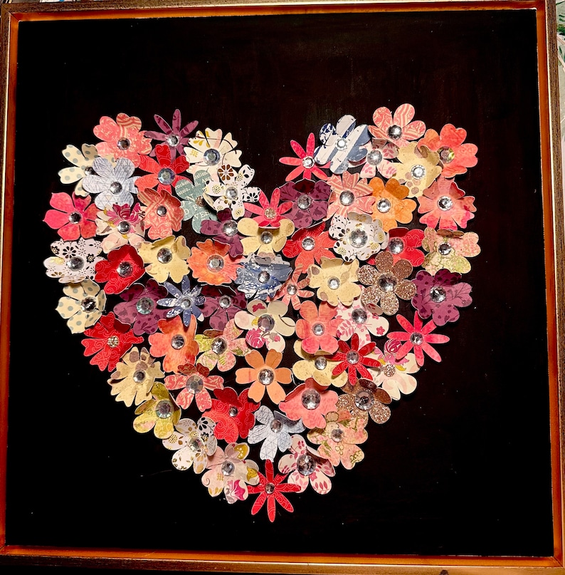 May include: A framed heart-shaped artwork made of colorful paper flowers with jeweled centers. The flowers are arranged in a heart shape on a black background.