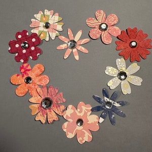 May include: A heart shape made of ten paper flowers with different patterns and colors. The flowers are arranged in a circle with the center of the circle open. The flowers are all different colors, including red, pink, blue, and green. Each flower has a small silver rhinestone in the center.