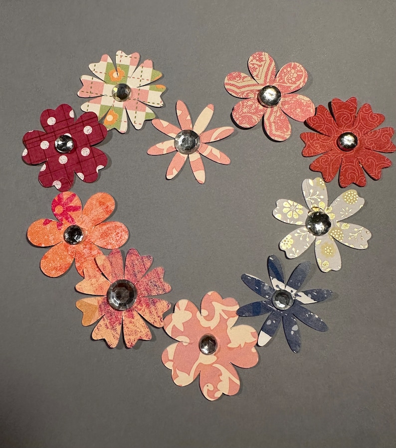May include: A heart shape made of ten paper flowers with different patterns and colors. The flowers are arranged in a circle with the center of the circle open. The flowers are all different colors, including red, pink, blue, and green. Each flower has a small silver rhinestone in the center.