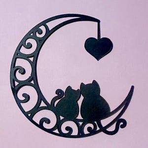 May include: A dark green metal wall art piece featuring two cat silhouettes sitting on a crescent moon. A heart hangs from the moon's curve. The moon has decorative swirls.