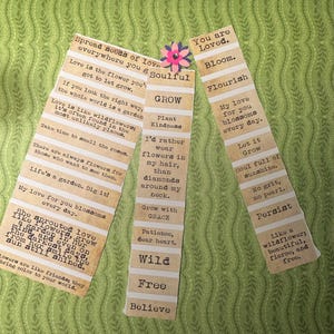 Puede incluir: Tres marcap&aacute;ginas de papel con citas inspiradoras. Los marcap&aacute;ginas tienen una est&eacute;tica vintage con un dise&ntilde;o a rayas beige y blanco. Una peque&ntilde;a flor de papel est&aacute; unida a un marcap&aacute;ginas. El fondo es un estampado floral verde.