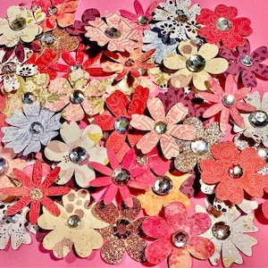 May include: A collection of paper flowers with rhinestones in the center. The flowers are in various shades of pink, red, and white. The flowers are arranged in a cluster on a pink background.