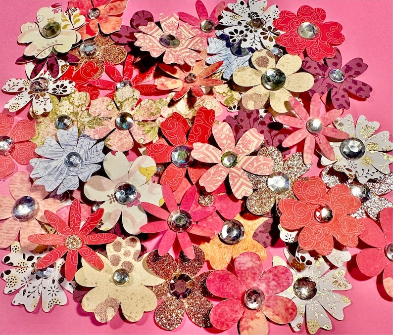 May include: A collection of paper flowers with rhinestones in the center. The flowers are in various shades of pink, red, and white. The flowers are arranged in a cluster on a pink background.
