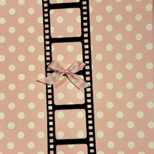 May include: A black film strip with five empty frames on a pink background with white polka dots. A pink and white gingham bow is tied around the film strip.