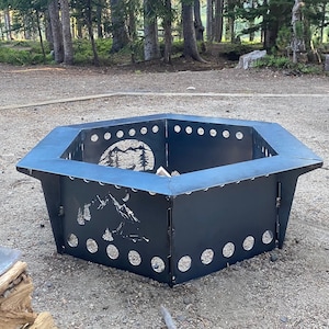 May include: A black metal hexagonal fire pit with a mountain and moon design. The fire pit is sitting on a gravel surface in a forest setting.