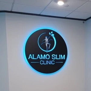 May include: A white leather couch with wooden legs sits in a waiting room with a black and blue logo that reads "Alamo Slim Clinic" on the wall. Two framed floral prints hang on the wall behind the couch.