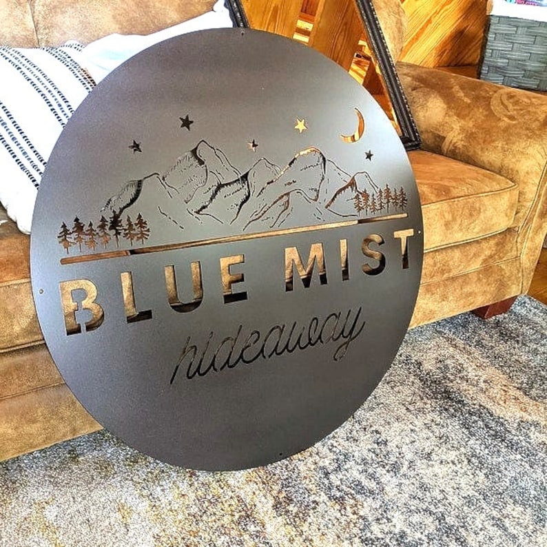 Business Sign, Logo, Large Outdoor Metal Wall Art, Custom Signs Business
 A round metal sign with a black background and gold lettering. The sign features a mountain range silhouette with stars and a crescent moon. The text on the sign reads 'BLUE MIST hideaway'.
