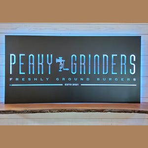 May include: A rectangular sign with the words "PEAKY GRINDERS" in a stylized font, with the tagline "FRESHLY GROUND BURGERS" and "ESTD 2021". The sign has a black background with blue neon lighting and is mounted on a wooden shelf.