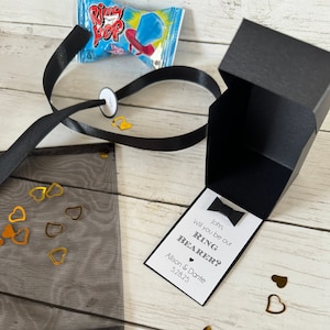 May include: A black box with a white card inside that says "John, will you be our Ring Bearer? Allison & Dante 5.28.25". The box is open and there is a black ribbon and a small bag of blue ring pops next to it. There are gold heart confetti scattered around the box.