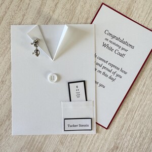May include: A white card designed to resemble a doctor's coat, with a button and a small silver pin. It includes a pocket with an eye chart and a name tag that reads "Tucker Stessis." A separate card with red trim reads "Congratulations on receiving your White Coat!"