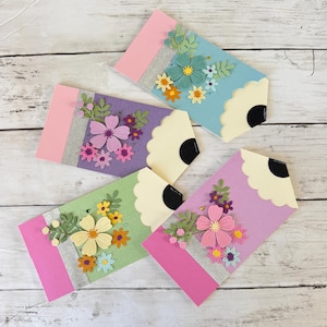May include: Four colorful pencil-shaped greeting cards with floral embellishments. Each card features a pink eraser, a silver band, and a decorative flower arrangement in shades of blue, purple, green, and pink. The pencil tips are cream-colored with a black center.