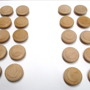 May include: A collection of twenty round, light-colored wooden knobs arranged in two vertical columns of ten. Each knob features a smooth, circular top surface with a subtle recessed ring detail, showcasing the natural grain and texture of the wood.