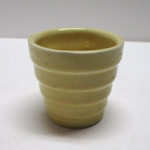 May include: A small, pale yellow ceramic planter with a ribbed design. The planter has a cylindrical shape with a slightly flared top. The exterior features horizontal ridges, adding texture to the piece. Ideal for small plants or succulents.