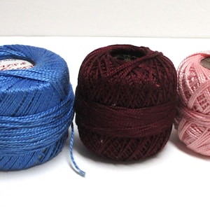 May include: Three spools of thread in blue, burgundy, and pink. The blue spool has a label that reads "Coats & Clark". The thread is tightly wound, and the spools are arranged side by side on a white surface. Ideal for sewing and crafting projects.