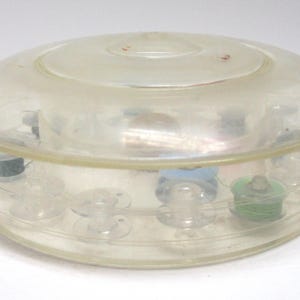 May include: A clear, round plastic sewing box with a domed lid. The box contains several spools of thread in various colors, including green and blue. The box is designed to store sewing supplies.
