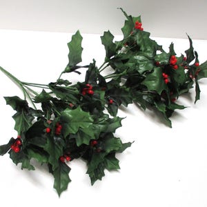 May include: An artificial holly branch featuring dark green, serrated leaves and clusters of small, bright red berries. The branch has a central stem that splits into multiple smaller stems, each adorned with foliage and berry groupings throughout.