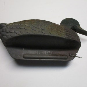 Puede incluir: Un se&ntilde;uelo de pato de pl&aacute;stico negro y verde con un dise&ntilde;o realista. El se&ntilde;uelo tiene un anillo de metal en la parte inferior y el texto "Clam-Dak" es visible en la parte inferior.