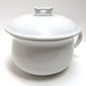 May include: A white ceramic chamber pot with a lid. The pot has a rounded body, a small handle, and a lid with a loop handle. The chamber pot is a vintage item.