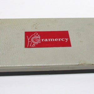 May include: A rectangular, light gray storage case with a textured surface and a dark base. Centered on the lid is a red rectangular label featuring a white graphic logo and the text Gramercy in white, sans-serif lettering.