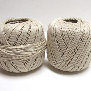 May include: Two beige yarn balls are displayed against a white background. The yarn is tightly wound, with visible strands creating a textured surface. The yarn appears to be a natural fiber, suitable for various crafting projects.