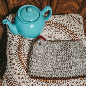 May include: A light blue teapot sits on a white crocheted doily with a gray and white fuzzy chicken-shaped hot water bottle.