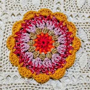 Può includere: Un sottobicchiere all'uncinetto fatto a mano con un motivo floreale. Il sottobicchiere presenta un vivace centro rosso e arancione, circondato da petali rosa, bianchi e beige. Il bordo esterno è smerlato, giallo dorato.