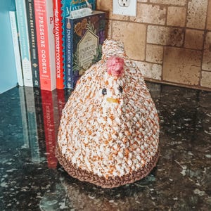 May include: A crocheted chicken pot holder in white and orange with brown accents. The chicken has a pink comb and a yellow beak.
