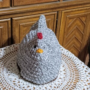 May include: A gray crocheted chicken with a red comb and yellow beak. The chicken is sitting on a white crocheted doily.