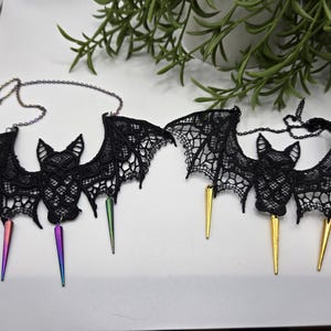 May include: Two black lace bat necklaces with chain accents. Each bat has intricate wing detailing and a pendant with a metallic spike. One necklace has rainbow spikes, the other has gold.
