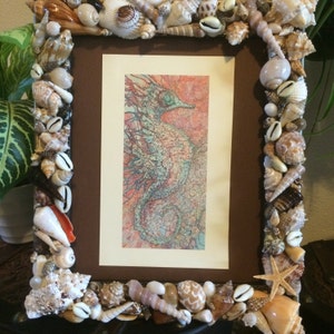 May include: A brown picture frame decorated with seashells, featuring a colourful illustration of a seahorse inside. The frame is made of wood and has a brown finish.