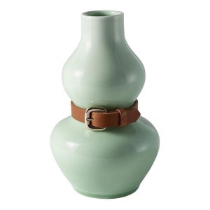 May include: A light green ceramic vase with a brown leather belt around the middle. The vase has a rounded, gourd-like shape.